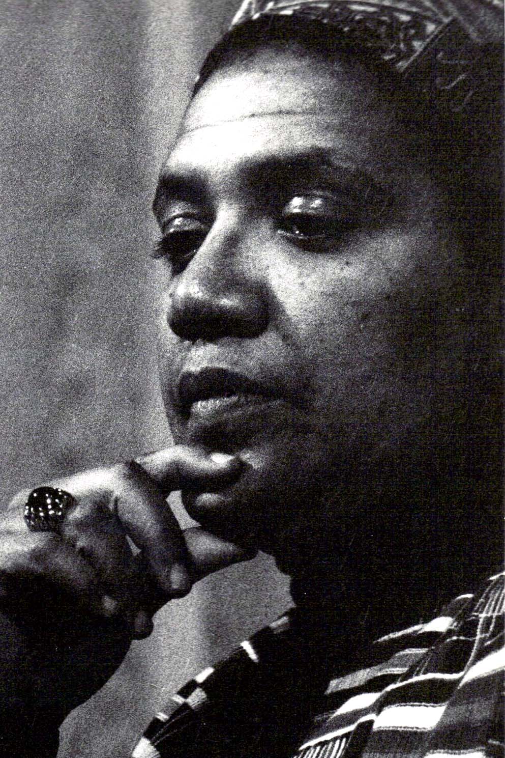 Closeup of Audre Lorde wearing a patterned turban, kente scarf, and jeweled ring and holding her chin with the thumb and forefinger of her right hand